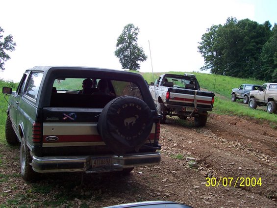 Rear view of a dark green Ford Bronco with a spare tire on the back, followed by a white Ford pickup truck and other SUVs on a dirt road