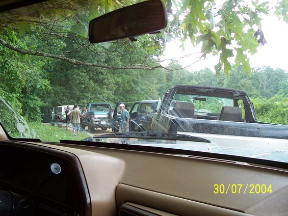 Several off-road vehicles parked on a dirt path in a wooded area, viewed from inside another vehicle