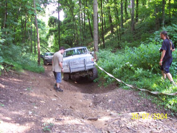 Trucks on a dirt trail in the woods, one being pulled by a strap. The front truck appears to be a 1980s Ford F-Series