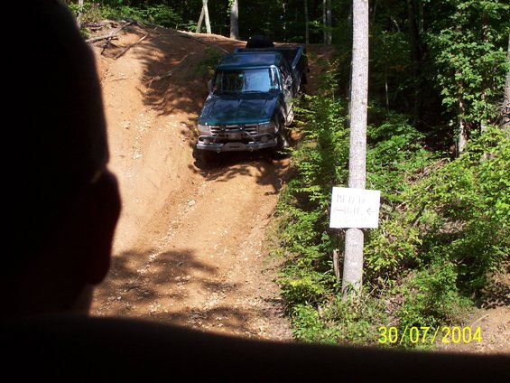 Green pickup truck driving up a steep dirt hill with trees on the side. Sign on a tree reads 'Muddy Hill'