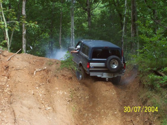 Dark blue Ford Bronco climbing a dirt hill, kicking up dust. Spare tire mounted on rear door
