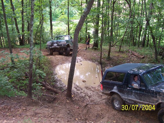 Two off-road trucks navigate a muddy path with a large puddle in a wooded area. A date stamp reads 30/07/2004