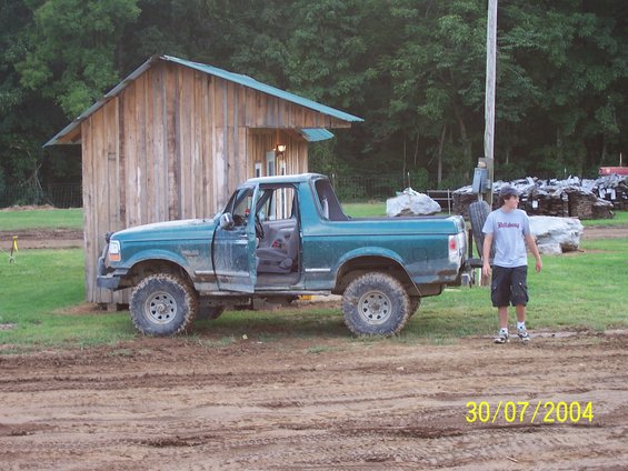 Green Ford Bronco with doors open, parked on dirt in front of a wooden building. A person stands to the right