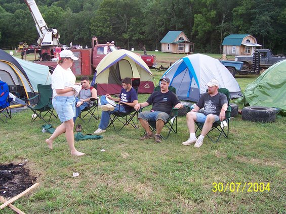 Campground scene with people sitting in chairs near tents, a red truck with a crane, and other vehicles in the background