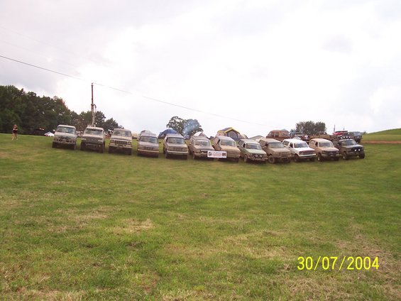 Multiple off-road trucks and SUVs parked in a line on a grassy hill with tents in the background