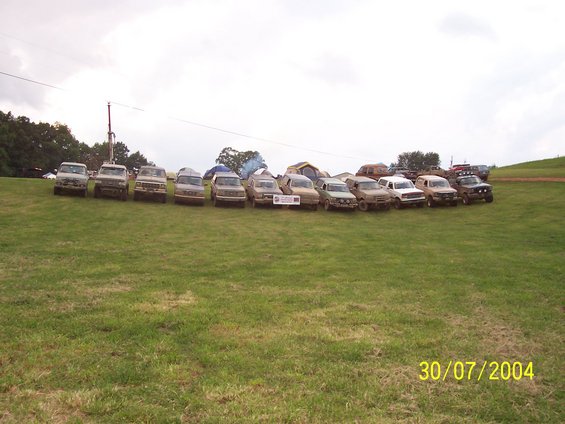Line of pickup trucks and SUVs parked on a grassy field with tents in the background