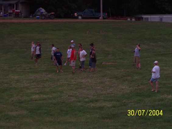 Group of people gathered in a grassy field with a building and vehicles in the background