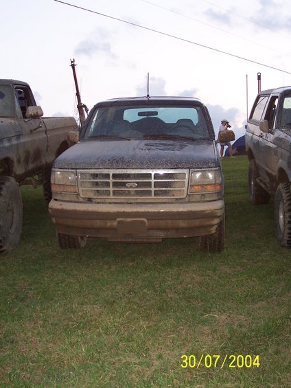 Mud-covered black Ford truck, front view. Grille has horizontal bars and a central Ford emblem. Headlights are rectangular. Tires are large and off-road style