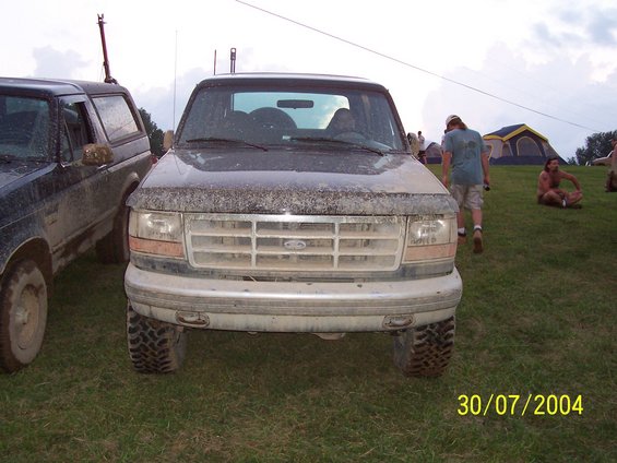 Front view of a dirty Ford truck parked next to another SUV. People and tents are in the background