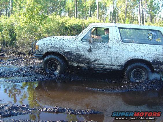 White Ford Bronco covered in mud driving through a muddy trail