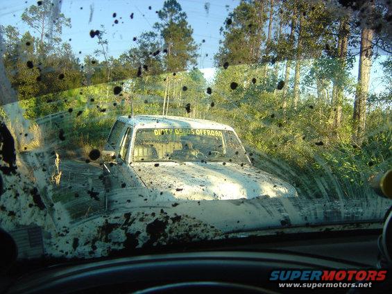 Mud-covered pickup truck with 'DIRTY DEEDS OFFROAD' sticker on windshield, stuck in mud with trees in background