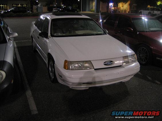 White sedan parked at night. It has a sunroof and a Ford badge on the grille. A red car is parked next to it