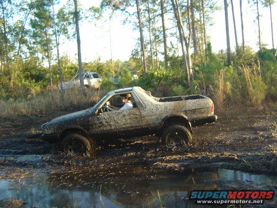 Mud-covered pickup truck with a person inside driving through a muddy puddle in a wooded area