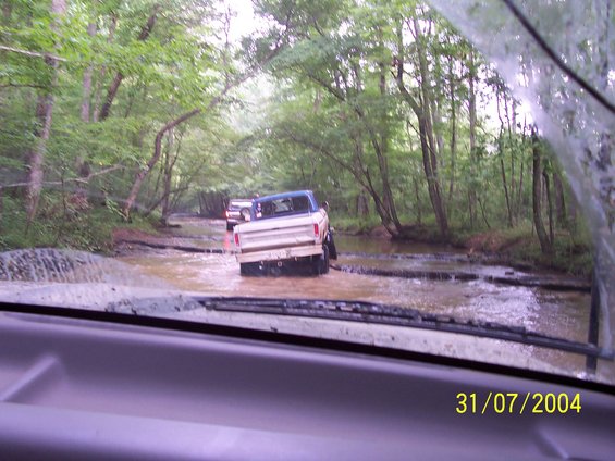 Two trucks driving through a muddy stream in a wooded area. The lead truck is a 1979 Ford F-150
