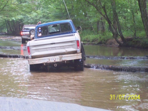 Silver and blue 1973 Ford F-150 truck driving through shallow water, rear view