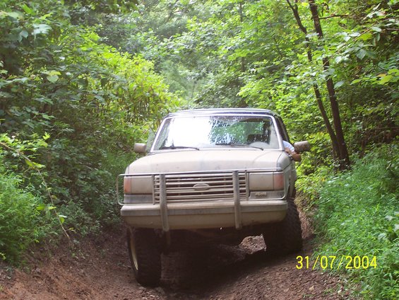Ford truck with a chrome grille and brush guard driving on a muddy dirt road through trees