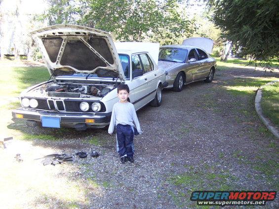Kid standing in front of a white sedan with its hood open, another car behind it