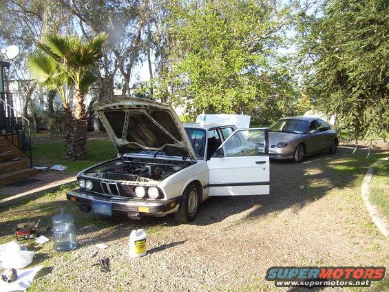 White BMW E30 sedan with hood and driver door open, parked on gravel. Another car is behind it