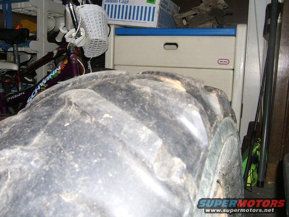 Close-up of a large, knobby off-road tire. A bicycle is visible behind it