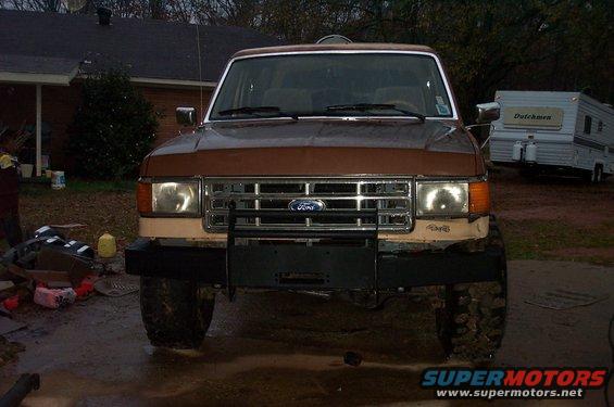Front view of a brown 1987 Ford Bronco with a new bumper and grille. A trailer is parked behind it