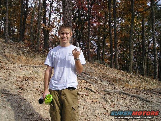 Young man in a white t-shirt and cargo shorts stands in a wooded area, holding a green water bottle and a remote control