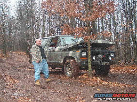 Man in camouflage jacket and jeans stands by a 1978 Ford Bronco with hood open, parked on a muddy forest road