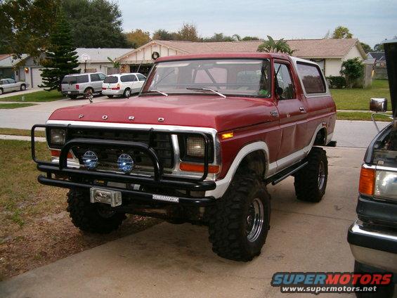 Red Ford Bronco with large tires, winch, and brush guard. Another vehicle is partially visible to the right