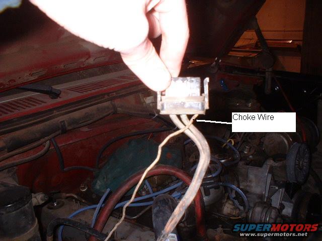 Hand holding a connector labeled 'Choke Wire' in a vehicle engine bay