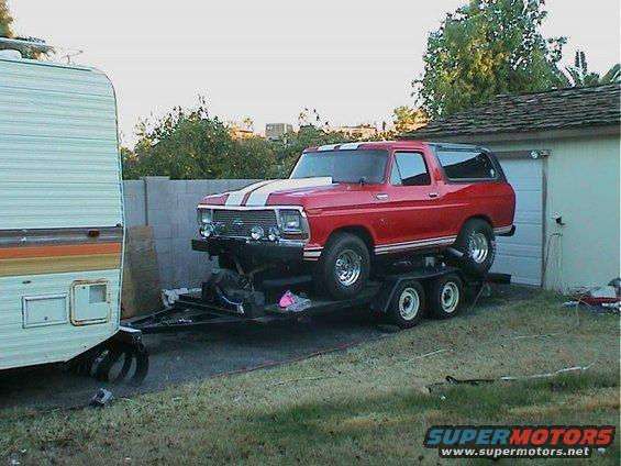Red Ford Bronco with white stripes on a trailer next to a camper and a garage