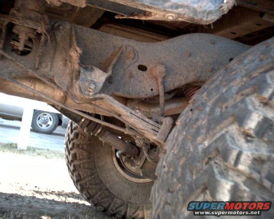 Underside of a dirty vehicle showing leaf springs, shock absorber, and a large tire. Another vehicle is partially visible in the background