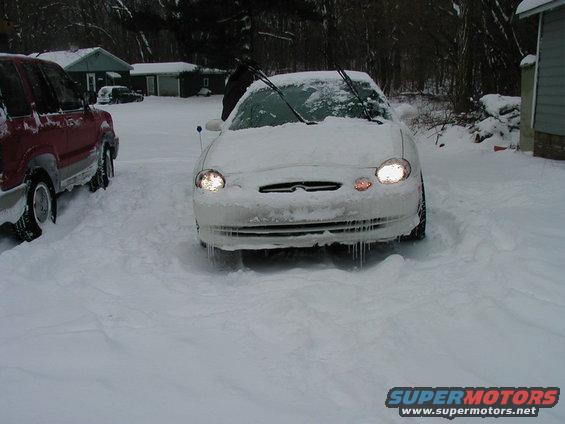 Front view of a white sedan in snow, icicles hanging from the bumper. A red SUV is parked to the left