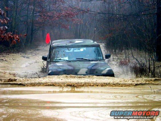 Camouflaged SUV driving through a muddy, wooded area