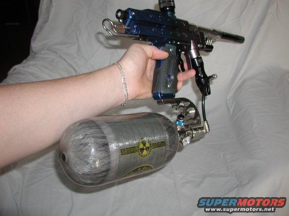 Hand holding a blue and silver paintball gun connected to a large grey air tank with a yellow radioactive symbol