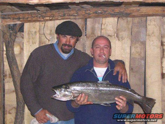 Two men pose for a photo holding a large trout. One man wears a flat cap and sweater, the other wears a jacket and has a cigarette in his mouth. A watermark reads SUPERMOTORS.NET
