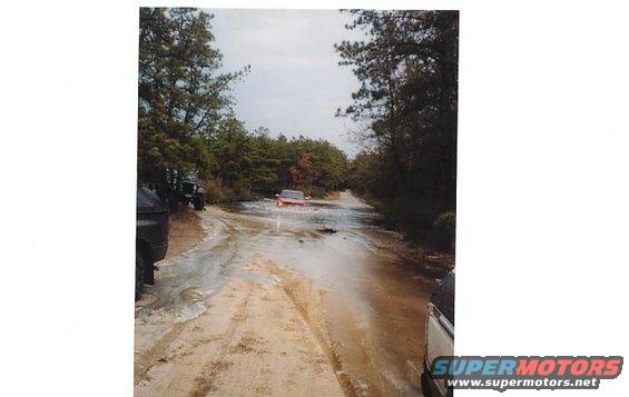 Red SUV driving through a muddy, flooded dirt road with trees on either side. Another vehicle is partially visible on the right