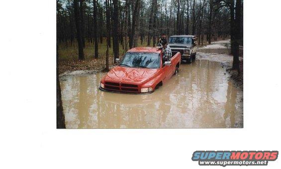 Red pickup truck stuck in mud, another truck behind it in a wooded area