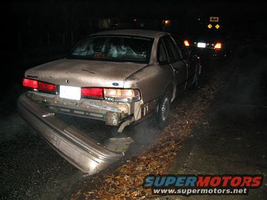 Rear of tan 1990s Ford Crown Victoria, bumper detached, parked on roadside at night