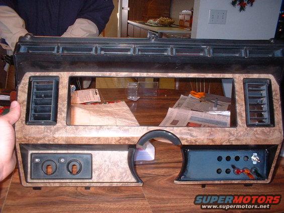 Dashboard trim panel with wood grain finish, air vents, and switch cutouts