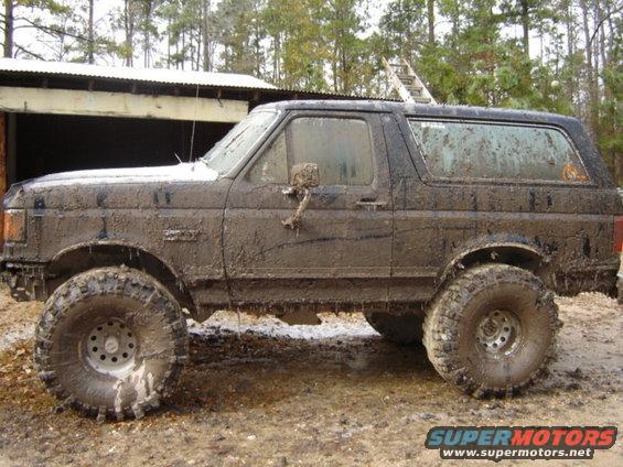 1989 Ford Bronco covered in mud, parked on muddy ground near a shed