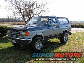Two-tone blue and silver 1987 Ford Bronco parked on a grassy lawn with a tree behind it