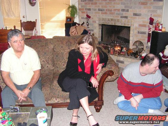 Three people are in a living room with a fireplace and couch. Christmas decorations are visible