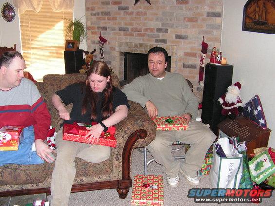 Three people sitting on a couch and floor, opening Christmas presents in a living room