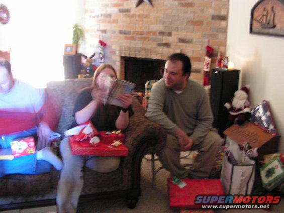 Three people sitting on a couch and chair, opening Christmas presents in a living room
