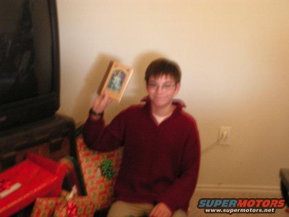 Boy in maroon sweater and glasses holds up a framed picture, with gifts and a TV visible