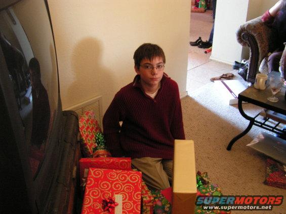 Boy in maroon sweater sits by a large black object, surrounded by wrapped gifts
