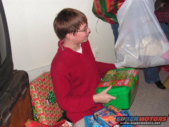 Kid in red sweater holding a green toy box, surrounded by wrapped presents