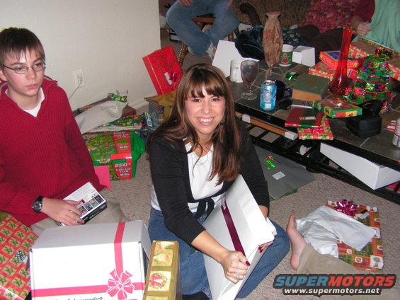 People opening Christmas gifts indoors, with a watermark reading SUPERMOTORS.COM
