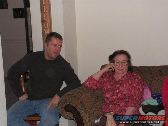 Man in dark shirt and jeans points while sitting next to woman in floral shirt on couch. "WELCOME" sign visible