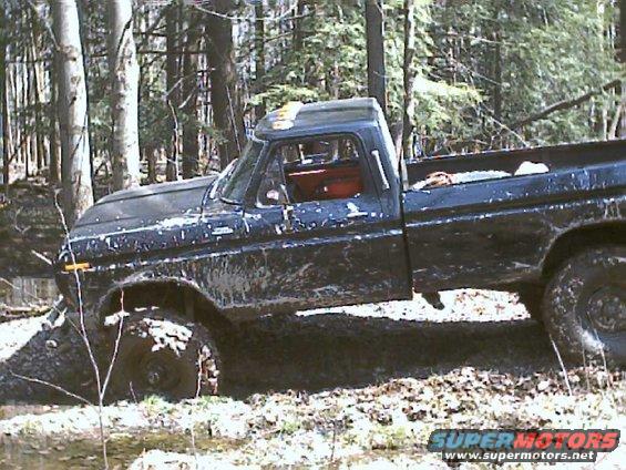 Black Ford pickup truck stuck in mud with trees around. The truck appears to be from the late 1970s
