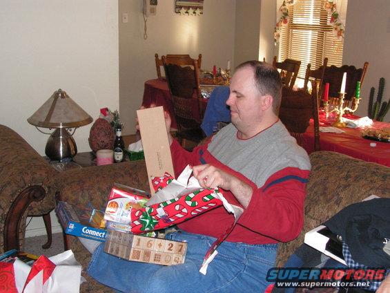 Man sitting on a couch opening Christmas presents. There are several wrapped gifts and a game box in front of him
